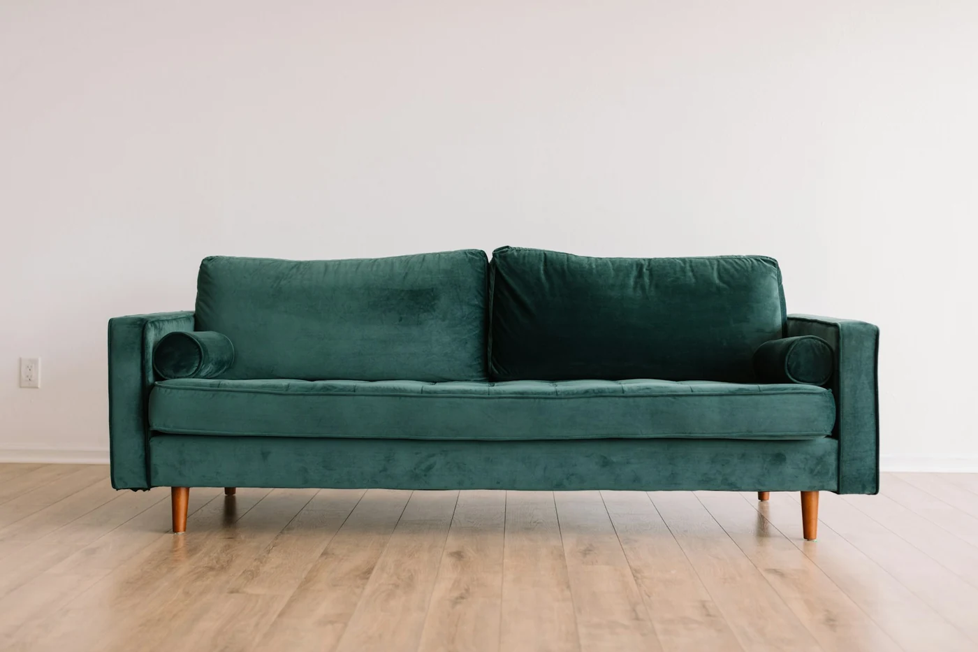 Zielona welurowa sofa trzyosobowa na drewnianych nóżkach pod białą ścianą