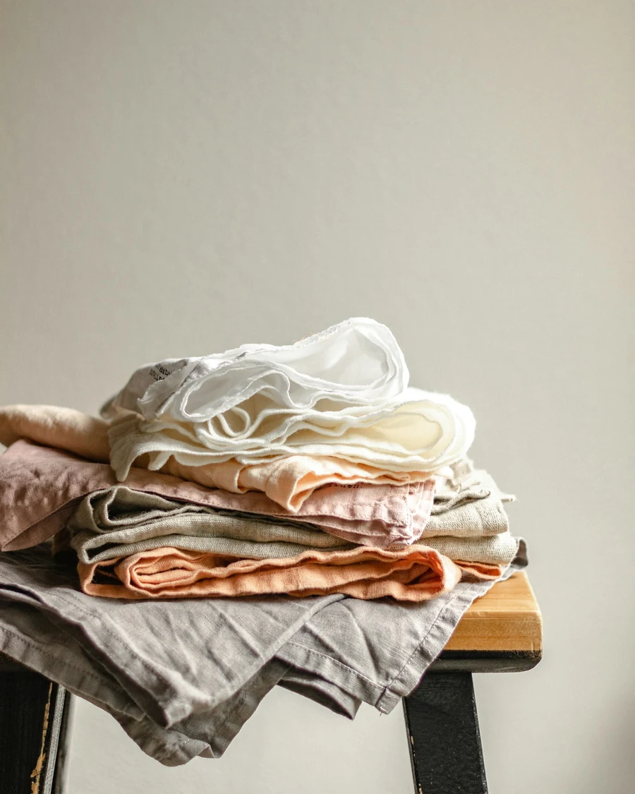 A stack of folded linen garments in white, peach and grey on a wooden stool