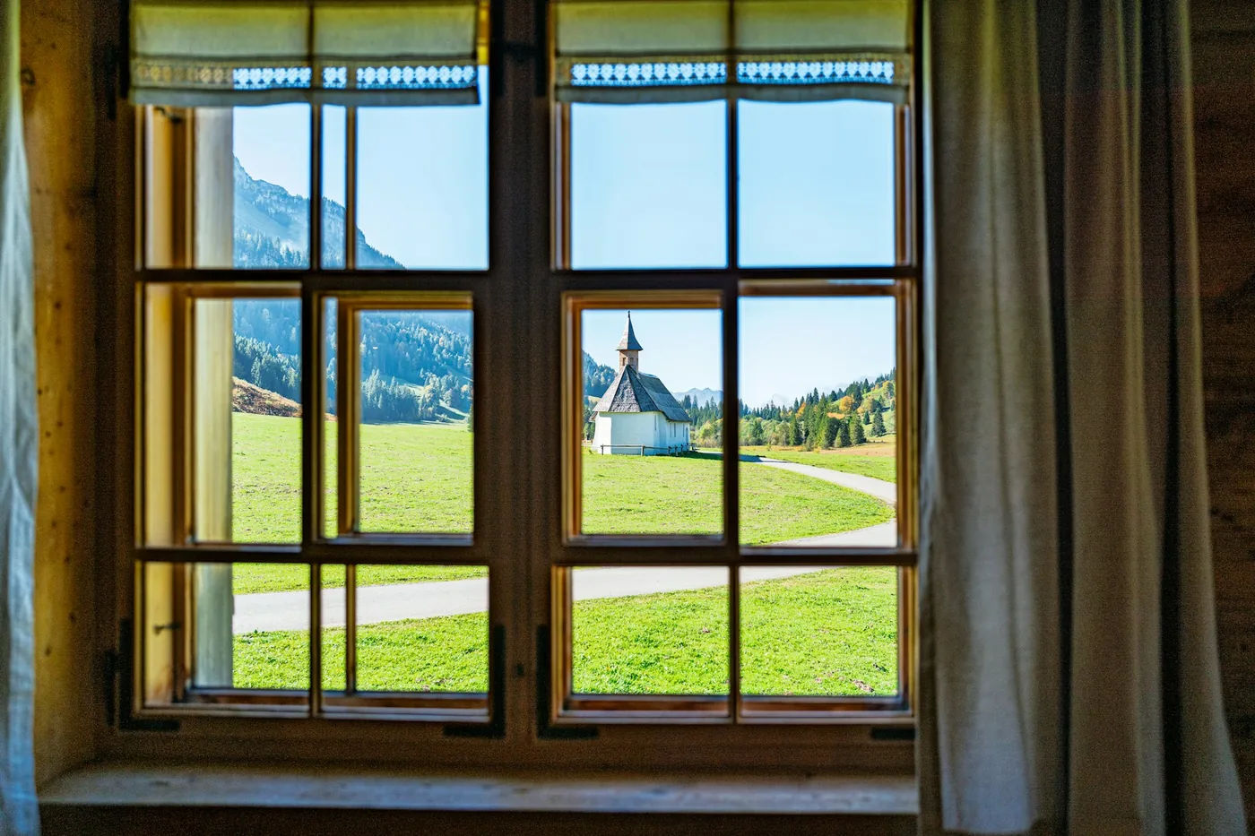Blick aus einem Holzfenster auf eine grüne Wiese und eine kleine Kirche