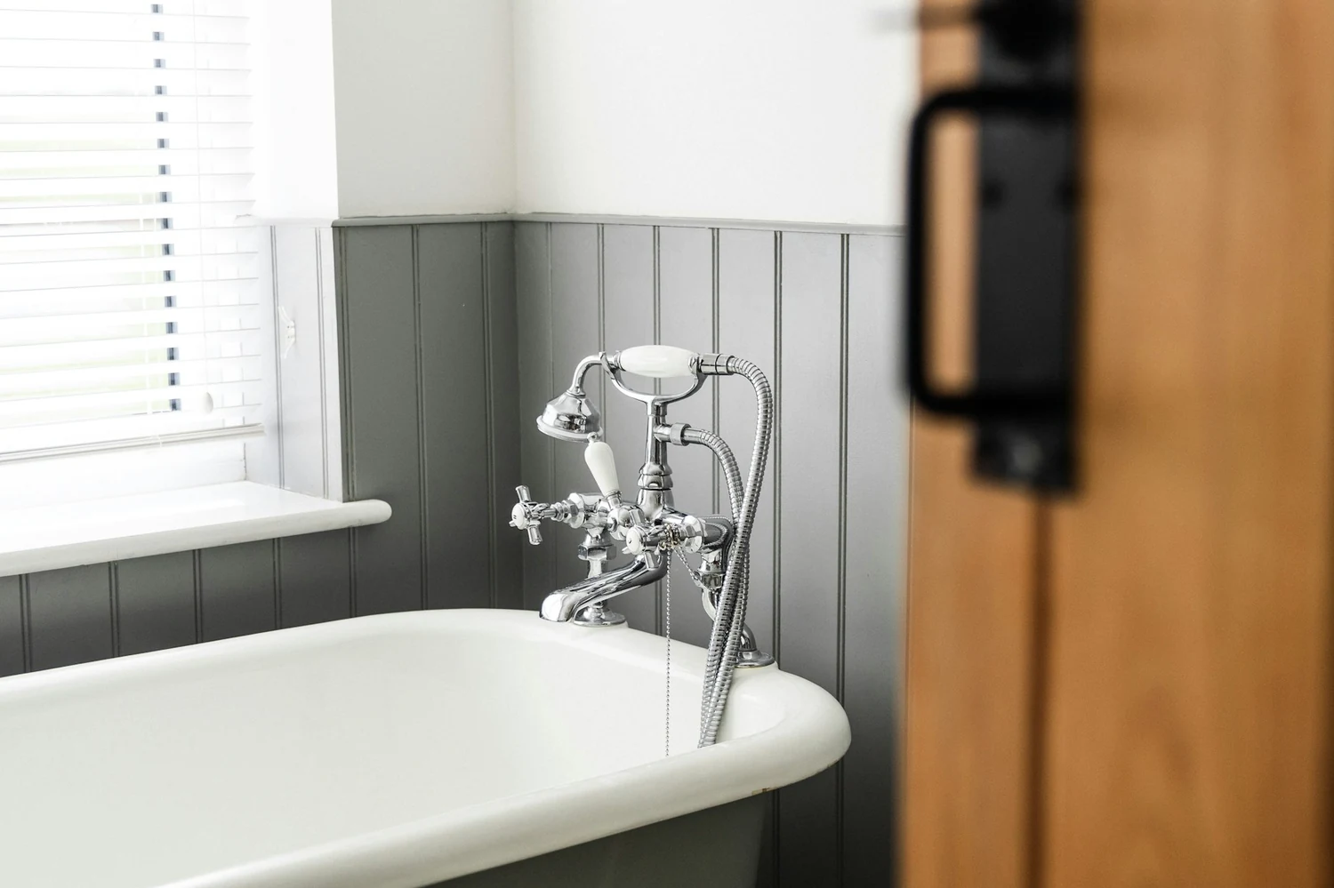 Weiße Badewanne auf Füßen mit verchromter Armatur am Fenster, Holztür