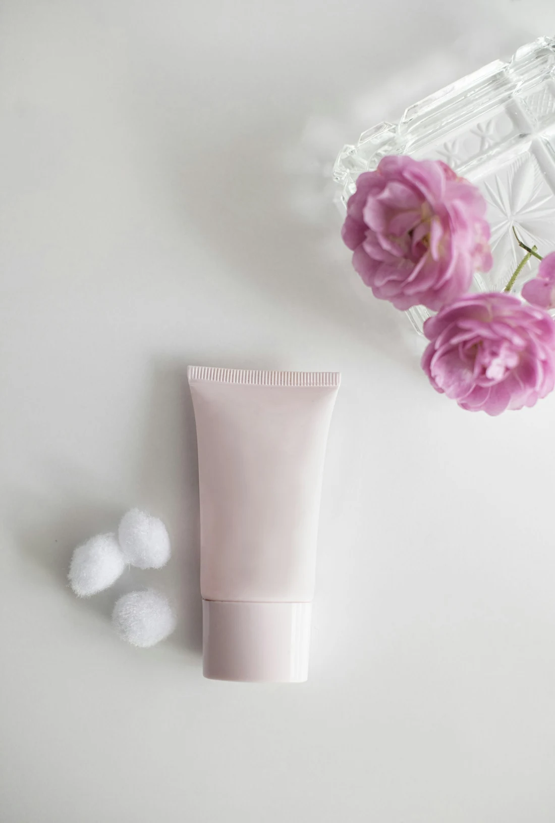 Tubo de crema blanco rosado junto a unas flores rosas y bolitas de algodón
