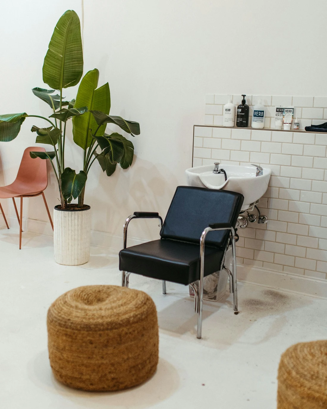 Rincón del salón con un sillón de lavado negro, azulejos blancos, un puf de esparto y plantas